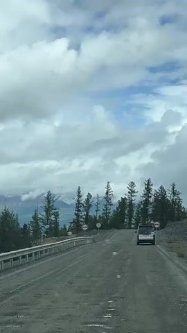 A black SUV is driving on a gravel road in a mountainous, forested area. The road is lined with a metal guardrail on the left and sparse trees on the right. Several road signs are visible, including "no passing" signs. The sky is partly cloudy with patches of blue. The overall scene suggests a remote, outdoor environment.