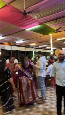 The video shows a gathering of people inside a large tented space, likely for a celebration or event. The ceiling is covered with alternating panels of pink, yellow, and green fabric. Several women dressed in saris move about the space, some possibly dancing, while others are seated at tables. A tall decorative light fixture stands near the center of the space. People walk around, some in casual attire, others in more formal wear. The event appears lively and social.