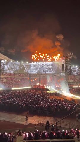 The video begins with a wide shot of a nighttime outdoor stadium concert. A large crowd is visible, holding up illuminated light sticks that create a sea of light. Fireworks are exploding behind the stage, where performers are visible on large screens. The camera then focuses on a woman in the crowd who is holding a light stick. The light stick changes color and shines brightly on her face as she smiles and seems excited. Other people with light sticks can be seen around her. The video ends with the woman sticking her tongue out with the light stick illuminated in red.