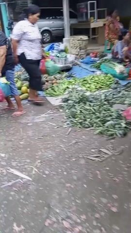 The video shows an outdoor market scene. Several people are visible, some walking and some standing. There are large piles of fruits and vegetables, including pineapples, papayas, and leafy greens, laid out on blue tarps. Some of the people appear to be shoppers or vendors. One woman in a striped shirt is walking past a display of pineapples. Another woman in a black shirt is carrying a blue plastic bag and a shopping bag. In the background, there are structures that look like market stalls and a van. The ground appears to be a mix of dirt and pavement.