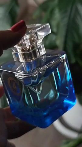 A hand with red fingernails holds a clear glass perfume bottle with blue liquid. The bottle has a silver cap and a circular emblem with the letter "A" on it. The background is blurred and green, suggesting foliage. The bottle is tilted and the light catches the liquid, creating reflections.