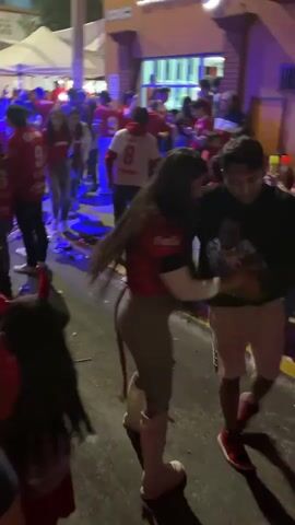 The video shows a lively street celebration at night. The crowd, mostly dressed in red, is dense, suggesting a large turnout. Many individuals are wearing red Toluca jerseys, and red and white flags are being waved in the air. The street is cluttered with debris, indicating the event has been going on for some time. The background reveals buildings illuminated with red lights, contributing to the overall festive atmosphere. People are walking, dancing, and interacting with each other, demonstrating the celebratory nature of the gathering.