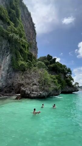 A drone shot captures a scenic view of a tropical bay. Lush green cliffs rise dramatically from the turquoise water, dotted with trees and vegetation. In the foreground, four people are swimming and playing in the clear, shallow sea. The sky above is a vibrant blue, with scattered white clouds drifting by. The overall impression is one of a peaceful and idyllic vacation spot.