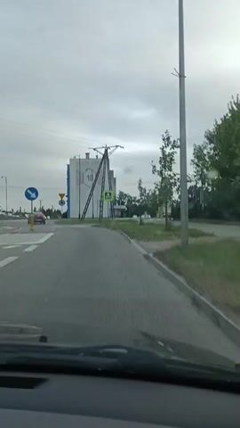 A vehicle is driving on a cloudy day. The vehicle is approaching a roundabout. There are power lines and a tall pole with wires overhead. There is a white building with a large hexagonal design on it. There are traffic signs, including a pedestrian crossing sign. A car passes by in the opposite direction. The roundabout has a red and white striped border. Another car is visible in the distance. A Polish flag is flying on a flagpole. There is a tall, brick industrial chimney in the background. Another building is visible behind it. More trees are present. The road has a pedestrian crossing marked on it. The sky is overcast.