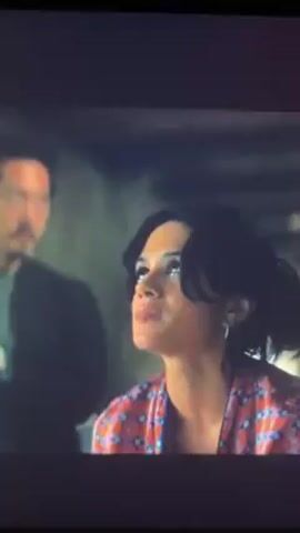 The video opens with a woman in a red and white patterned shirt talking. A man with a mustache stands in the background. The scene cuts to two men standing in a mostly empty room with blue walls. The room has a niche with a small statue inside. The man on the left is wearing a black jacket and black pants. The man on the right is wearing a black hoodie and blue jeans. The man on the left turns to look at the man on the right.