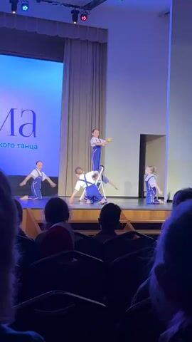 The video shows a dance performance taking place on a stage in front of an audience. Five young girls are performing. They are wearing matching outfits consisting of white shirts and blue overalls with white straps. The backdrop is a large screen displaying a blue background with white writing. One girl stands on a white step ladder holding a yellow object, while the others dance around her. Some girls perform cartwheels, while others pose with their arms raised. The audience is visible in the foreground, seated in rows of chairs. The stage has wooden edges, and there are closed beige curtains on either side of the stage screen. The lighting is bright and focuses on the stage.