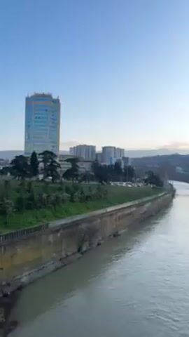 The video shows a cityscape viewed from a bridge over a wide, grey river. Buildings of varying heights, including several high-rises, line the riverbanks. Construction cranes are visible among the buildings on the right bank. Trees, largely bare of leaves, are present along the riverbanks, as is a low wall. The sky is pale blue, with the sun setting or rising near the horizon on the right side of the frame, partly obscured by clouds. The water in the river is moving swiftly. There are reflections of the sky and buildings on the water's surface.