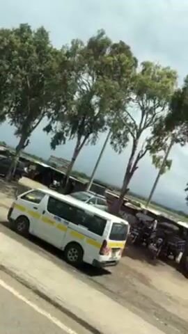 The video shows an African roadside market from a moving vehicle. The camera is tilted slightly, and there are trees and vehicles visible on the left of the road. A white van with yellow and green stripes passes in the foreground.  The market appears to have small stalls and vendors. The sky is overcast.