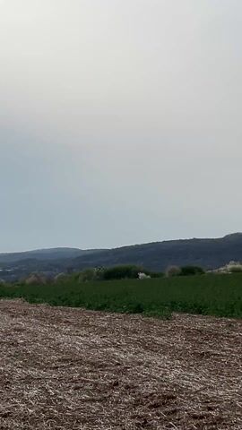 The video shows a landscape with a field in the foreground and a wooded hill in the background. The field appears to be partially tilled and partially covered in green plants. A light gray sky dominates the scene. Towards the middle of the video, a crop duster airplane appears, spraying something over the field.