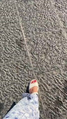 This media file captures a first-person perspective of someone walking on a cobblestone street in Catania, Italy. The scene is set outdoors, bathed in bright sunlight, suggesting a clear and warm day. The subject is wearing casual attire, featuring a pair of light-colored slip-on sandals with red-painted toenails, and flowing pants with a blue and white floral pattern. The focus is on the lower half of the body and the ground beneath, with the textured cobblestones filling most of the frame. A prominent shadow cast by the walker extends across the stones, indicating the time of day is likely midday or early afternoon due to the angle of the sun. The overall impression is one of leisurely movement through a charming urban environment, characteristic of Italian city streets. No visible text is present in the media.