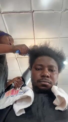 A person with dark, curly hair is getting their hair styled. A second person, wearing an apron, is using a blue comb and a blow dryer on the first person's hair. The first person is looking directly at the camera. There are white towels draped around their neck. The ceiling appears to be a drop ceiling with fluorescent lights.