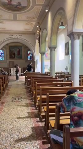The video pans across the interior of a church. Pews are arranged in rows on either side of a central aisle. The aisle is carpeted with a patterned runner. In the foreground, a person wearing a brightly colored shirt is seated in a pew, facing away from the camera. Further down the aisle, several people are standing and walking, some appearing to be tourists. The church walls are decorated with religious artwork and stained-glass windows. The ceiling is adorned with ornate paintings and molding. The lighting is natural and artificial, creating a serene atmosphere. The overall impression is of a peaceful, sacred space being visited by individuals.