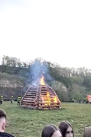 The video captures a large bonfire constructed from stacked logs, burning in a grassy field. A figure, resembling a witch effigy, is positioned atop the log pile and is engulfed in flames, producing considerable smoke. The fire is active and vibrant at the base of the structure.

Several individuals in yellow high-visibility vests, likely firefighters or event staff, are visible standing at a safe distance around the bonfire. A white safety barrier or tape delineates the immediate area around the fire. In the background, a wooded hill with a dense tree line rises against an overcast evening sky.

In the foreground, several spectators are gathered, watching the event unfold. The faces of two young women are partially visible in the bottom right, with one holding a smartphone displaying what appears to be a social media interface or video call.

The scene suggests a communal gathering, likely an observance of a traditional event such as Walpurgis Night or "pálení čarodějnic" (witch burning) common in Central Europe, where bonfires and effigies are burned. The location is Dalovice, Czechia, and the time is evening, as indicated by the ambient light and the prominent glow of the fire.