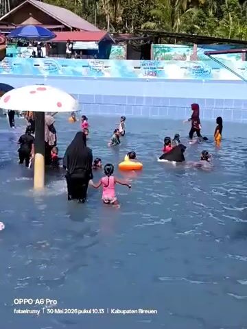 The video shows a group of people, including women wearing hijabs and children, enjoying a swimming pool at what appears to be a water park. The pool has light blue water. A mushroom-shaped structure stands in the middle of the pool, spraying water. In the background, there is a water slide and other structures, suggesting a recreational area.  The weather appears sunny and clear. The video has a text overlay in the bottom left corner indicating the device used to record it: "OPPO A6 Pro". It also provides a timestamp: "30 Mei 2026 pukul 10.13 Kabupaten Bireuen," suggesting the video was taken on May 30, 2026, at 10:13 AM in Bireuen Regency.