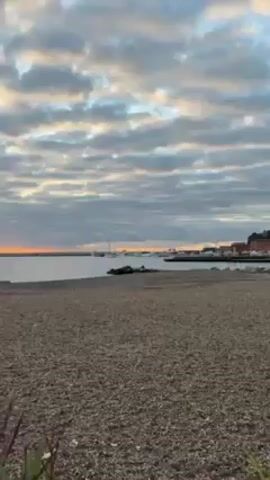 The video shows a beach with small pebbles at dawn. The sky is covered with many clouds, and the rising sun creates an orange glow. In the distance, land masses can be seen over the sea, with a port on the right-hand side. A person with a dog appears in silhouette on the beach, walking and interacting with each other. There are pieces of driftwood on the beach.