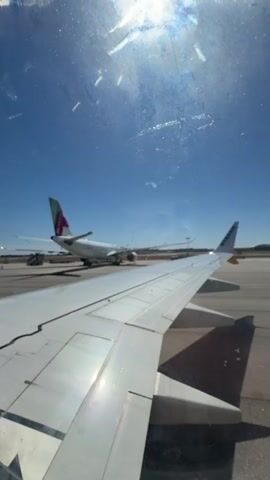 The video shows a view from inside an airplane, looking out the window at the tarmac. The wing of the airplane is visible in the foreground, and another airplane is parked in the background. The sky is clear and blue, and the sun is shining. The video appears to have been taken during the daytime, likely at an airport.