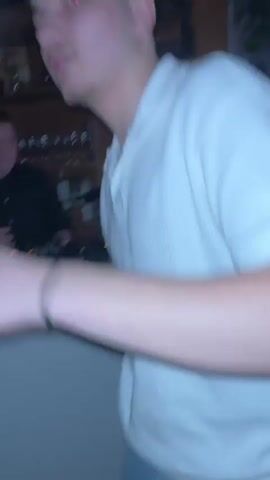 Here's a detailed description of the video:

The video shows the interior of a dimly lit space, possibly a small bar or club. The scene is somewhat shaky and blurry due to camera movement. A person, presumably a DJ, is visible in the background, wearing a black shirt and standing behind a DJ console with illuminated buttons and knobs. There is some greenery visible to the left of the frame.

In the foreground, there are people, their faces partially obscured or out of focus. Some of them raise their arms, appearing to be dancing or enjoying the music. The lighting creates a warm, slightly orange hue in parts of the background. The environment has a rustic or woody feel, possibly due to wood paneling on the walls. There are also glasses on a shelf in the background.
