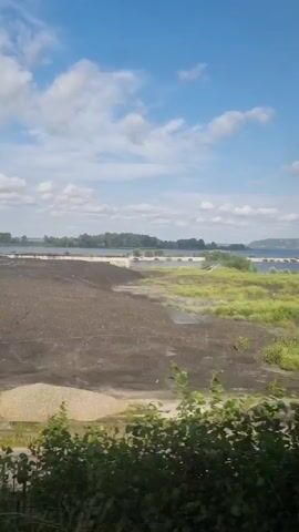 The video captures a dynamic outdoor scene, likely from a moving vehicle, showcasing a wide expanse of landscape under a bright, partly cloudy sky. The foreground is dominated by a dark, rough terrain, possibly industrial slag heaps, interspersed with patches of vibrant green grass and scrubby bushes. Beyond this, a large body of water, likely a river or lake, stretches across the midground. On the opposite bank of the water, a line of trees forms a natural horizon, with distant structures, possibly buildings or bridges, visible against the horizon.

The scene appears to be set during the daytime, with the sunlight casting distinct shadows and illuminating the landscape. The weather seems pleasant, with a clear blue sky dotted with fluffy white clouds, suggesting a warm, perhaps summer day. The presence of lush greenery and the general ambiance evoke a sense of nature reclaiming an industrial area, or a transitional zone between human activity and the natural world.

There are no people or easily identifiable objects like vehicles or animals that are the primary focus. The activity taking place is implied by the slight camera movement, suggesting travel through or past this location. The location cues, combined with the visual elements, align with descriptions of the Trypillia area in Ukraine, which is known for its historical significance and its proximity to the Dnieper River. The overall emotion conveyed is one of observation, a fleeting glimpse of a landscape that blends the remnants of industry with the resilience of nature. No visible text is present in the video.