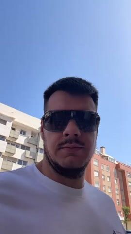 The video begins with a close-up shot of a man wearing sunglasses and a white t-shirt, standing outdoors. The background shows a blue sky and buildings. The man brings a paper cup to his lips and takes a sip of a beverage. He then lowers the cup and makes a facial expression. The scene appears to be a casual, outdoor setting on a sunny day.