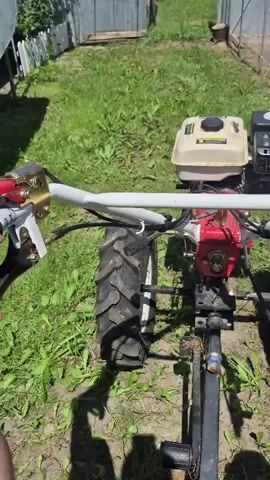 A person's hand is shown holding the throttle of a red and white walk-behind tractor. The tractor has large, treaded tires and a small engine on the back. The scene appears to be outdoors in a grassy yard with a wooden fence in the background. The sun is shining, casting shadows on the ground. The tractor is not moving.