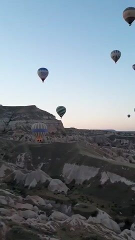 A drone shot captures a panoramic view of a mountainous landscape at dawn. Numerous hot air balloons, illuminated from within, drift gracefully across the clear, pale blue sky. The balloons are of various colors and patterns, some featuring stripes, others solid colors, and one with text. The landscape below is rugged, characterized by distinctive rock formations and valleys, with patches of green vegetation. The sun has not yet fully risen, casting a soft light on the scene.