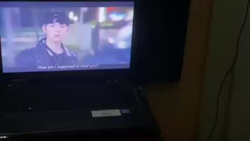 Here's a detailed description of the video:

A laptop is positioned in front of a larger display screen, which is wall-mounted. The laptop is black with an HP logo visible. On the laptop screen, which is open, a Korean drama with English subtitles is playing. The drama shows different scenes of characters talking to each other. The visible text from the subtitles includes, "How am I supposed to trust you?" and "Aren't you leaving? You were in a hurry." The display screen behind the laptop shows the same content as the laptop screen, just at a larger scale. The background of the setting appears to be a room with a wooden wall. The lighting is dim.