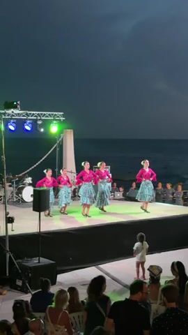 This video captures a lively outdoor performance at Es Camp De Mar, Spain. A group of five young women, dressed in vibrant pink tops and turquoise patterned skirts, are performing a synchronized dance on a raised stage. Their costumes suggest a cultural or festive event, possibly a luau or folk dance performance. The stage is illuminated by theatrical lighting, with red, blue, and green spotlights casting colorful hues on the dancers and the surrounding area. To the left of the stage, a drum set and speaker are visible, indicating a live musical accompaniment.

The scene is set against the backdrop of the ocean at dusk. The sky is a deep twilight blue, suggesting the event is taking place in the evening. The calm sea is visible behind the stage, and the gentle waves hint at a pleasant coastal atmosphere. A crowd of spectators is seated in front of the stage, their faces turned towards the performers, some holding up phones to capture the moment. The overall mood appears to be celebratory and engaging, with the audience enjoying the vibrant performance. A young child in the foreground is also watching the show, adding to the family-friendly atmosphere. The overall setting is a beachside promenade or plaza, typical of a resort town in Spain. No visible text is present in the video.