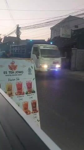 The video shows a roadside scene at dusk or dawn. In the foreground, a portion of a white car is visible. To the left, a large sign displays various flavors of "Es Teh Jowo" (Javanese Iced Tea) with images of the drinks and prices. The drinks appear to be fruit-flavored teas with toppings. In the background, a two-lane road is visible with several businesses and buildings lining it. A motorcycle with a rider is passing by, and a large white truck is also on the road. Overhead, numerous power lines are strung across the sky. The lighting suggests it is either early morning or late evening.