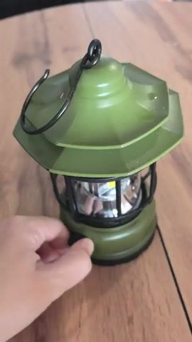 A hand is shown turning a knob on the side of a green lantern, which is illuminated. The lantern is sitting on a wooden surface.