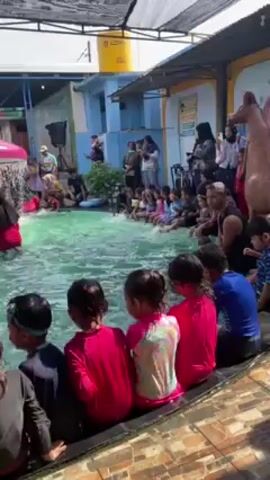 A group of children in swimsuits are sitting around the edge of a pool. Some are wading and splashing in the pool, which has a mushroom shaped water feature in the center. Adults are standing around the pool as well, some are taking pictures. The pool is outdoors in a recreational area.