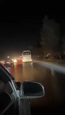 The video shows a night scene on a road with moderate traffic. The view is from inside a car, looking out the windshield. A large white bus is in front, with its taillights illuminated. Other cars can be seen in the distance. The road appears wet, reflecting the lights. Street lights illuminate the road. The sky is dark and clear. The car is moving slowly in traffic.
