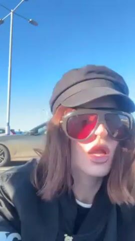 A person wearing a black cap, large red-tinted sunglasses, and a black jacket with a "CDG" logo is speaking to the camera. The person has reddish-brown hair. They say "I feel like I'm incognito." They are standing in a parking lot under a clear blue sky. The person holds a silver-colored bag in their left hand. They are wearing black pants with white stripes on the sides. The person is wearing black boots. Cars are visible in the background.