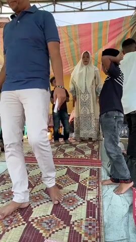 In an outdoor setting under a striped tarpaulin, a group of people are gathered on woven mats. A woman wearing a light-colored hijab and a patterned dress stands centrally, facing the camera. To her right, a boy in a dark t-shirt and jeans, wearing a black cap, stands with his back to the camera, his right hand raised towards the woman. To the woman's left, a younger boy in a brown t-shirt and jeans stands with his hands clasped in front of him. Further to the left, a man in a blue shirt and white pants is partially visible, with his back to the camera. Other individuals are visible in the background, including a man in a white shirt to the right of the woman and a person lying down on a mat in the far left background. The overall atmosphere suggests a casual gathering or event.