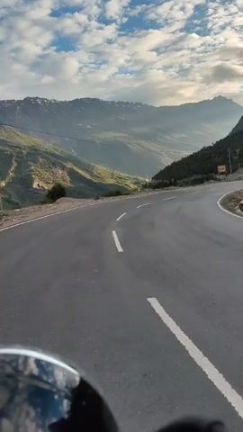The video shows a first-person perspective of someone riding a motorcycle or scooter on a winding mountain road. The road is asphalt with white lane markings. To the left of the road, there is a steep mountain slope covered in greenery and rocks, with a valley visible in the distance. To the right, the mountain rises sharply, with a few trees and rocks on its face. The sky is partly cloudy with a mix of blue sky and white clouds. The overall scene depicts a scenic drive through a mountainous landscape.