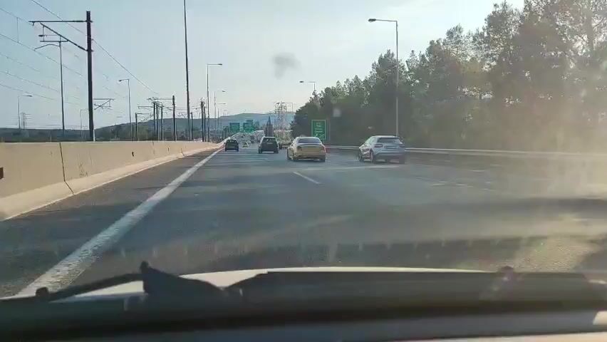 This video shows a dashcam view from a car driving on a highway near Koropi, Greece. The scene is set on a multi-lane highway with clear skies and bright sunshine, suggesting it is daytime, likely late afternoon due to the warm light.

The primary subjects are the vehicles on the road. Several cars are visible, including a yellow sedan, a silver SUV, and a dark SUV in front of the camera car. The highway itself is the main object of focus within the scene, with its asphalt surface, white lane markings, and a concrete barrier separating lanes. On the left side of the highway, there are overhead power lines and poles, typical of infrastructure found in many areas. In the distance, hills and mountains are visible, and some trees line the right side of the road. Green road signs with white text are present on overhead gantries, indicating upcoming exits or directions. One sign visible in the distance appears to have Greek lettering.

The activity is simple: driving on a highway. The cars are moving at a steady pace, and there are no indications of traffic jams or unusual events. The overall mood is that of a normal commute or travel. The weather appears clear and pleasant, contributing to a sense of ease. The presence of Greek text on the signs confirms the location as Greece, and the mention of Koropi in the prompt helps pinpoint the specific region. The image is clear, with no visible text other than the road signs.