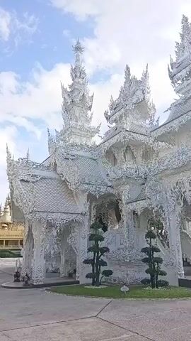 The video pans across a stunning, intricately decorated white temple. The architecture is ornate with numerous tiered roofs, spires, and detailed carvings covering the entire structure. Two small, manicured evergreen trees stand in front of the temple, flanking an entrance. The sky above is a mix of blue with scattered white clouds. The ground in front of the temple appears to be paved.