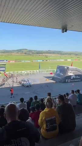 The video shows a live audience watching a motocross stunt performance. In the foreground, spectators are seated on bleachers, facing a wide grassy field under a bright blue sky. In the middle ground, a performer on a dirt bike is in mid-air, performing a stunt. The bike and rider are positioned above a ramp and an inflatable landing mat. A person in an orange vest stands near the ramp, possibly directing the performance or ensuring safety. In the background, rolling green hills are visible under a clear sky. The scene appears to be an outdoor event taking place on a sunny day.