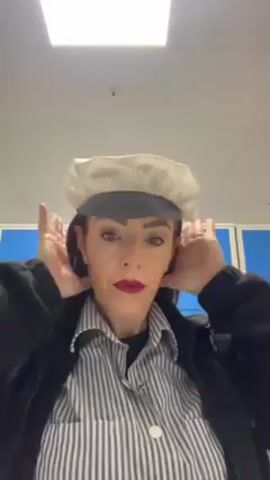 A woman stands in a room, likely a locker room, facing the camera. She wears a striped shirt, a black jacket, and a two-toned cap. Her hands are behind her head, possibly adjusting the cap or her hair. The background includes light-colored walls and blue lockers. The lighting is bright, possibly from fluorescent ceiling lights. She wears red lipstick and is looking directly at the camera.
