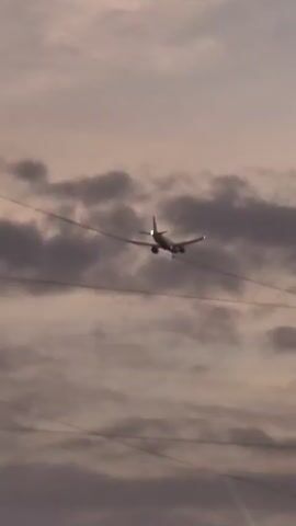 A plane is captured in mid-flight against a cloudy sky. The aircraft is angled downwards, suggesting it is either descending or approaching for a landing. Power lines stretch across the frame, appearing in the foreground and behind the plane. The lighting suggests it could be dusk or dawn, with soft, diffused light.