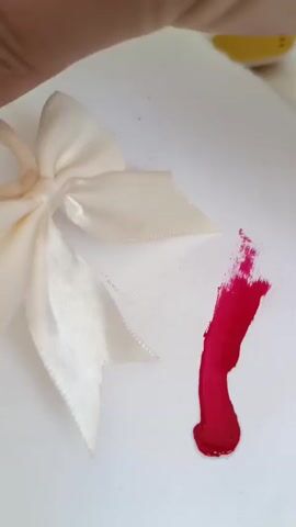 A close-up shot shows a person's fingers holding a light-colored ribbon bow with a loop. Next to the bow, a streak of bright red paint is applied to a white surface. The paint appears to be wet and is in a vertical stroke, with a thicker blob at the bottom. The lighting is bright, casting a slight shadow.