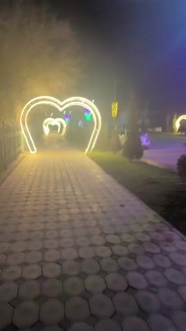 A low-angle, medium shot captures a paved path adorned with illuminated heart-shaped arches leading into the distance. The pavement is patterned with grey and white geometric shapes. On the left, a dark metal fence runs along the path, while on the right, a grassy area is interspersed with small trees. Beyond the path, a road is visible with parked cars, including a white van. In the distance, additional heart arches glow with a warm, soft light, and there are also some decorative lights in blue and green. The scene is shrouded in a light fog, which diffuses the light from the arches, creating a soft glow. The trees in the background are bare, suggesting it is either late autumn or winter.