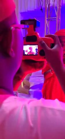 A fair-skinned person with brown hair, wearing a white shirt, is seen from the back. They are holding an orange camera with dark-skinned hands. The camera screen displays a person with dark skin, dressed in a red traditional African garment and matching headwrap, singing into a microphone. Another individual is visible in the background of the camera's display. The environment is lit with red and purple lights.