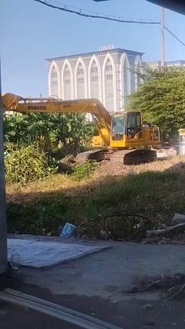 A yellow Komatsu excavator is parked on a patch of dirt and overgrown grass. In the background, a large building with arched windows and the word "UINSA" is visible. The sky is clear and blue.