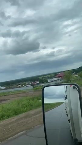 The video is shot from the perspective of someone in a vehicle, likely a truck, looking out the side window and in the side mirror. The scene outside the window shows a rural landscape with scattered houses, fences, green fields, and trees. The sky is overcast with heavy grey clouds. The side mirror reflects a similar view, with the road, sky, and a portion of the vehicle's exterior visible. The vehicle appears to be moving on a paved road that transitions to a dirt or gravel path. The overall impression is of driving through a somewhat cloudy, rural environment.