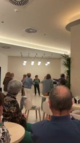 A group of older women are dancing in a circle in a brightly lit room while several older adults sit and watch. The dancers are dressed in casual clothing. The person at the front of the circle has an orange shirt, and is moving their arms more enthusiastically than the others. The observers sit at small tables with white chairs. A plant stands in the background.  A cluster of hanging lamps is visible above the dancers.
