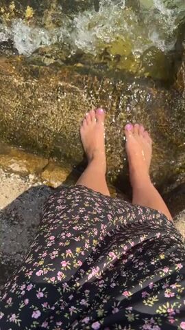 A person's feet are submerged in shallow, clear water. The person is wearing a dark floral print dress. The water is clear and shows rocks and sand on the bottom. In the background, there is a concrete dock with a boat tied to it. The water is calm and reflects the sky, which is blue with some clouds. Mountains are visible in the distance. The sun is shining, creating dappled light on the water and rocks.