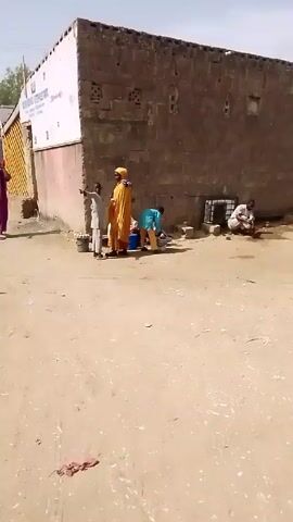 The video shows an outdoor scene in an arid area.  Several people are visible. One individual in a long, yellow garment stands near others who appear to be washing or preparing something using buckets and a low table.  Nearby, another person is seated on the ground and appears to be working. The camera moves, following a person in a long, patterned dress and a black headscarf. The scene shifts to a wider street view, showing people walking towards an entrance marked "FEDERAL UNIVERSITY DU". A car with its trunk open is parked to the side of the road.