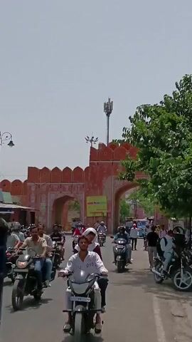 The video captures a street scene in Jaipur, India, with a prominent pink gate serving as a backdrop. The sky is overcast, and the scene is bathed in daylight. The focus is on the steady flow of traffic moving towards and through the gate. Most vehicles are motorcycles or scooters, with riders and occasionally passengers navigating the road. Pedestrians are also present, walking along the roadside. A large tree with lush green foliage stands to the right of the gate, partially framing the scene. A tall telecommunications tower rises in the background above the gate. The overall atmosphere suggests a busy urban setting with a mix of traditional architecture and modern elements.