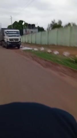 From a low first-person perspective, the video shows a truck and trailer passing on a paved road. The truck is white and pulls a grey trailer. The trailer has multiple axles. The road is reddish-brown. Along the side of the road is a light green wall, and some vegetation is visible. Further down the road, two people are walking. There are some potholes in the road.