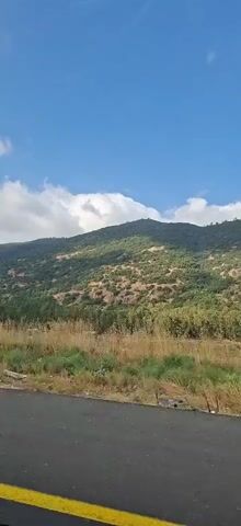 The video shows a view from a moving vehicle of a mountainous landscape. In the foreground, there is a paved road and dry grasses with some visible trash. The middle ground features a hillside covered in green trees and shrubs, with some patches of exposed earth. In the background, a large mountain rises against a blue sky with scattered white clouds. A tall, striped pole with a light fixture is visible on the left side of the screen at the beginning of the video. The overall impression is of a scenic, rural environment.